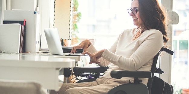 Glad kvinna med glasögon sittande i rullstol och använder en dator, foto