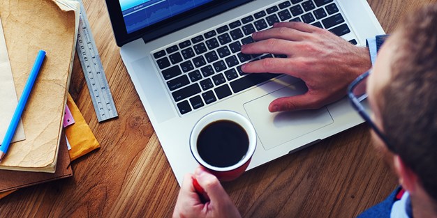 En man med ena handen på tangentbordet på sin laptop och den andra handen hållandes i en kopp kaffe. Foto