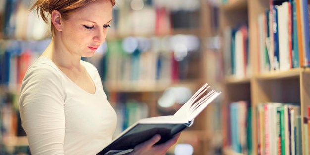 Ung kvinnlig student som bläddrar i en bok i ett bibliotek. Foto